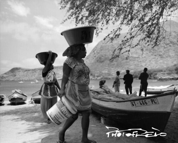 Deux femmes marchent sur la plage en portant des bassines sur la tête. À côté d'elles, posées sur la plage, il y a des barques de pêcheur.